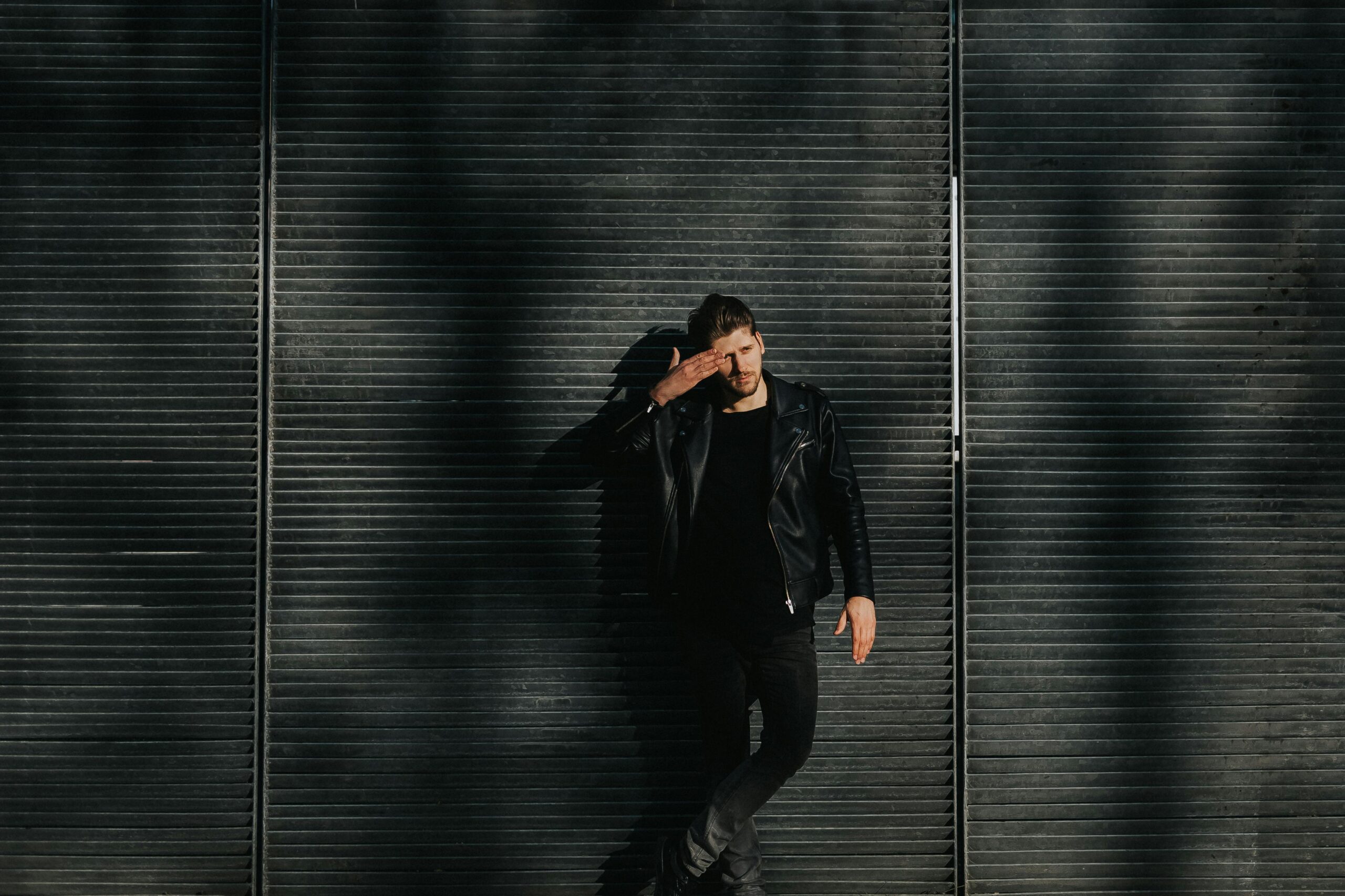 Young man poses confidently in a black leather jacket against a gridded metal urban wall.